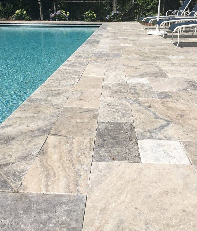 Light gray and beige pavers create a smooth patio surrounding a clear blue swimming pool, with poolside lounge chairs visible in the background.