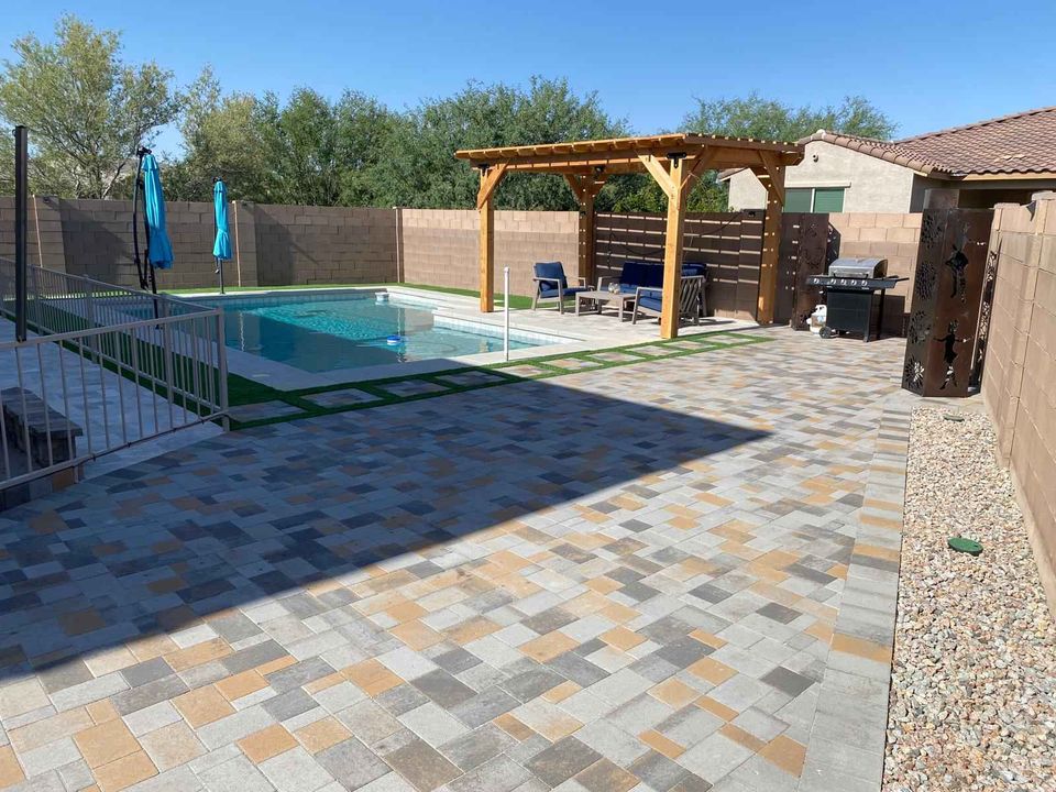 A fenced backyard pool area with a pergola, outdoor seating, grill, and umbrellas. The patio features pavers in multicolored tiles, while rocks line the edge near the brick wall and desert landscaping.