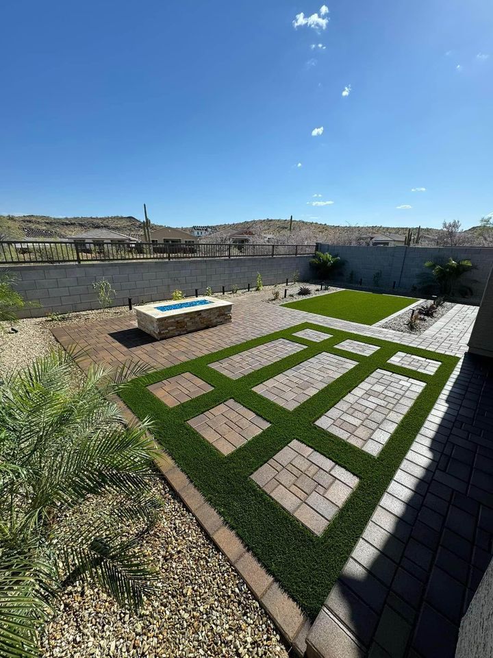 A modern backyard with geometric pavers set in artificial grass, a raised rectangular fire pit on a stone patio, and desert landscaping with rocks and drought-tolerant plants under a clear blue sky.