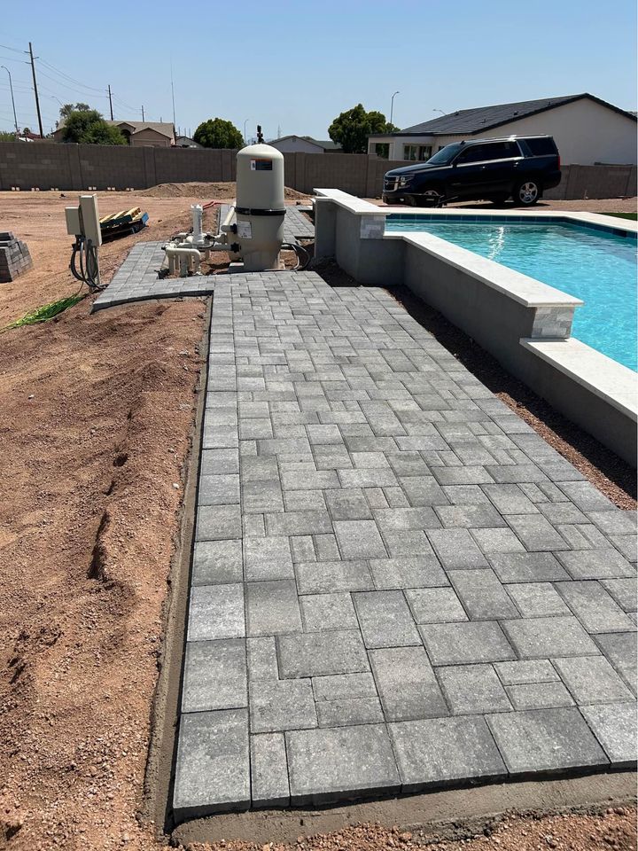 Gray pavers form a walkway beside a backyard swimming pool, with construction materials and equipment nearby, and a black SUV parked in the background under a clear sky.