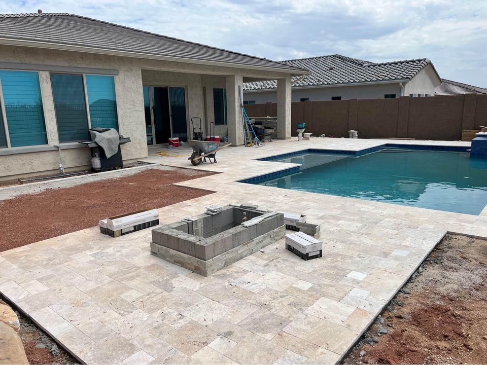 A backyard under construction with pavers forming a stone patio, an unfinished fire pit, stacks of bricks, and a swimming pool near a beige house. Tools and equipment are scattered around.