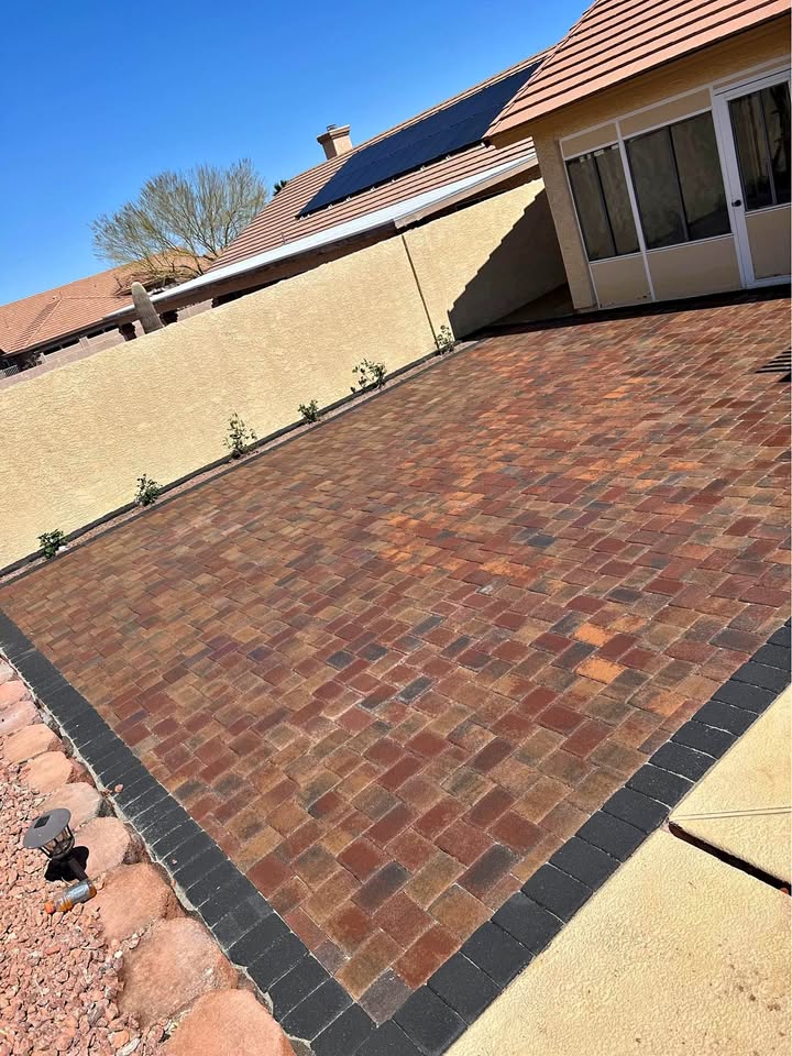 A backyard with a freshly installed brick paver patio bordered by black pavers sits beside a house with tan walls, a tiled roof, and solar panels under a clear blue sky. Small plants line the beige wall.