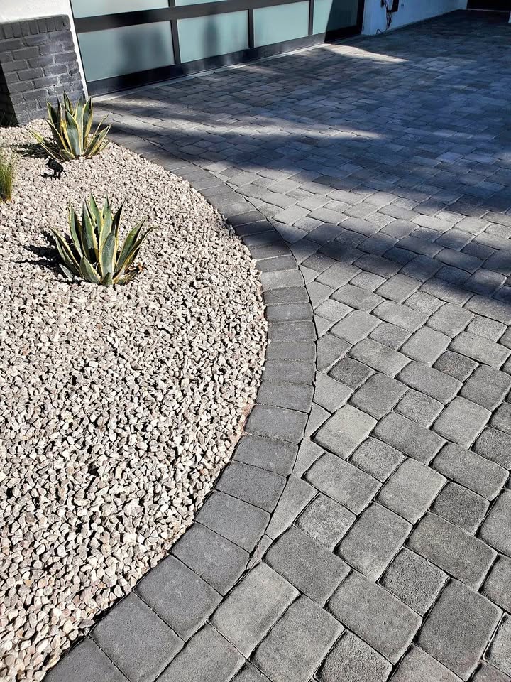 Curved pavers create a stylish driveway beside a landscaped area with light-colored gravel and two agave plants. The clean, modern space is complemented by a glass-paneled garage door in the background.