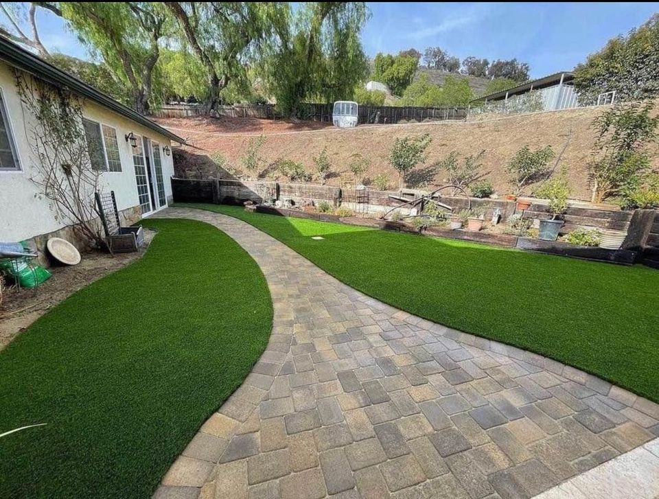 A backyard with a curved stone paver pathway, bordered by artificial green grass, leads past garden beds and a white house; terraced hillside with trees and plants in the background.