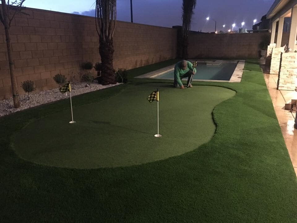A person installs or adjusts artificial grass for a small putting green with two golf flags in a backyard next to a swimming pool in the evening.