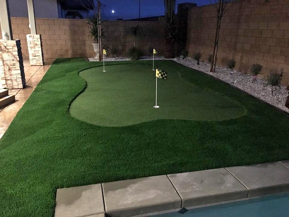 A backyard putting green with artificial turf features three holes, each marked with a yellow checkered flag. The green is bordered by landscaping rocks, plants, and a stone patio next to a swimming pool.