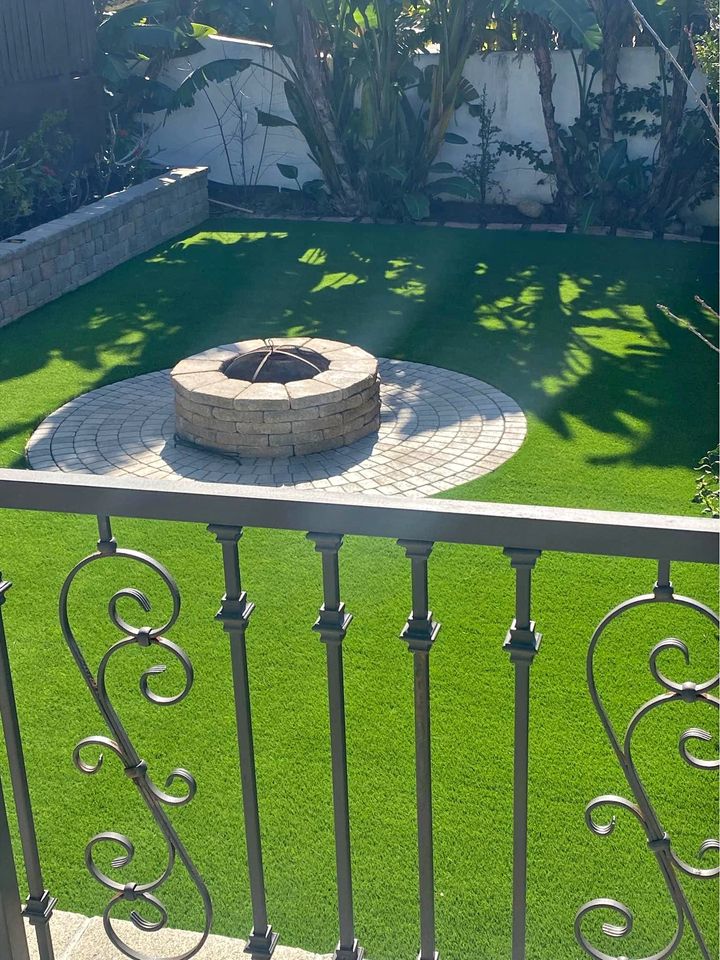A backyard with bright green artificial grass, a circular stone fire pit on a round paved area, and decorative black metal railing in the foreground. Lush plants and a white wall are in the background.