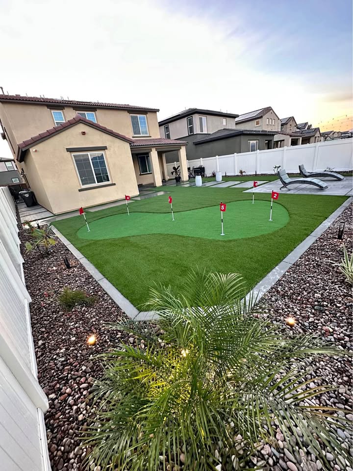 Backyard with artificial turf putting green featuring six holes with red flags, surrounded by rocks, plants, and a lounge chair, with a beige two-story house in the background.