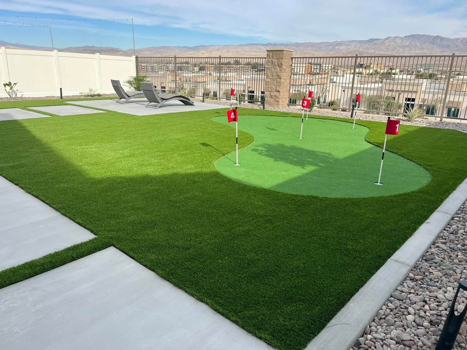 A backyard with artificial grass features a small putting green with four red flags, lounge chairs on a concrete patio, and desert mountains in the background under a clear sky.