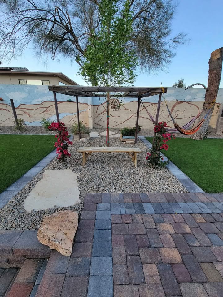 A backyard patio features a stone path leading to a wooden bench under a pergola with blooming pink flowers. There’s a hammock on the right, a tree in the center, and lush Elite Synthetic Grass on both sides.
