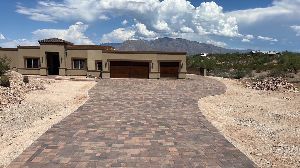A modern single-story house with a large brick-paved driveway, featuring Elite Synthetic Grass landscaping amid a desert setting, set against distant mountains and a partly cloudy sky.