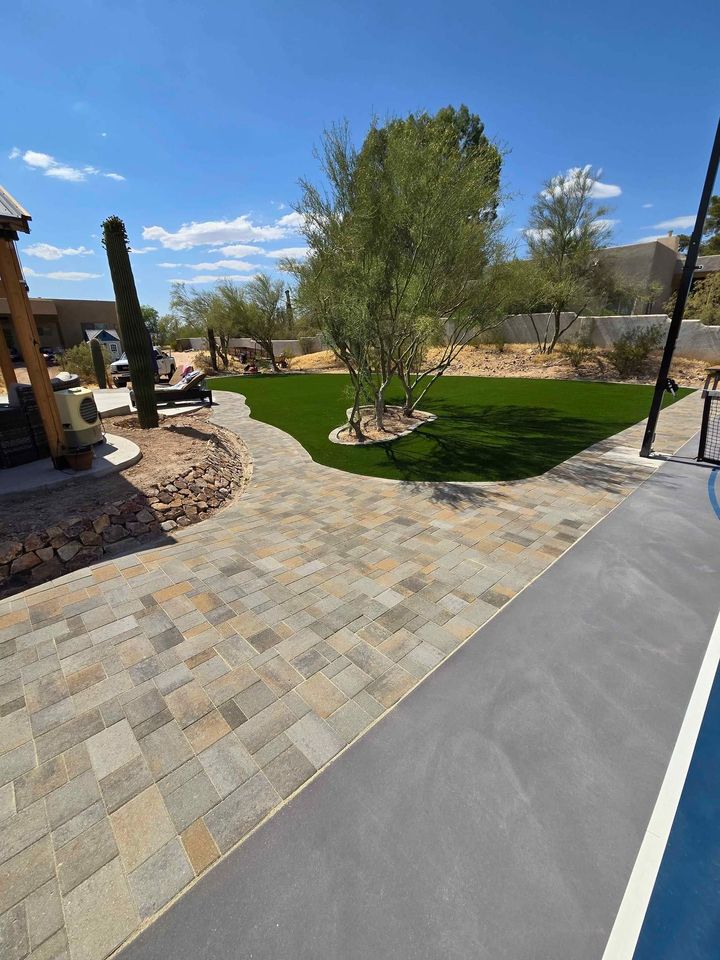 A backyard with a paved patio, Elite Synthetic Grass area, desert landscaping with rocks, and several trees and cacti under a clear blue sky.
