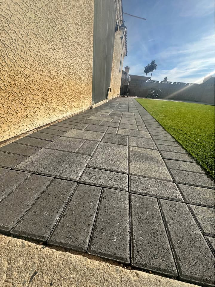 A paved walkway made of rectangular stone tiles runs alongside a tan stucco house, with Elite Synthetic Grass on the right and a sunny, clear sky in the background.